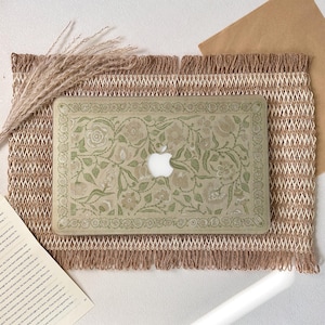 May include: A laptop with a floral design case featuring a white Apple logo. The case is light green with a pattern of flowers and leaves. It rests on a woven mat with a fringed edge, with dried plants and a book in the background.