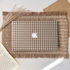 Op de afbeelding: Een bruin en wit geruit laptophoesje met het Apple-logo. De laptop staat op een getextureerde placemat met franjes. Gedroogde tarwearen en een stuk papier staan ook in de afbeelding.