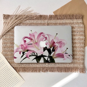 Peut inclure: Une coque rigide pour ordinateur portable arborant un motif imprimé de fleurs de lys rose clair avec des tiges et des feuilles vertes. La coque est transparente, laissant apparaître le logo Apple argenté au centre du motif floral.