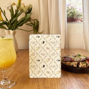 Peut inclure: Une coque de tablette crème avec un motif répété de chiens de dessins animés lisant des livres et des vignes vertes. L'étui est posé sur une table en bois. Un verre de boisson jaune et une pizza sont également sur la table.