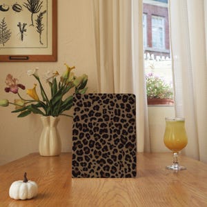 Puede incluir: Una funda rectangular para tableta con un diseño de estampado de leopardo en tonos marrón y negro. La funda está sobre una mesa de madera, junto a un jarrón con flores y un vaso de zumo. La escena está iluminada por una ventana.