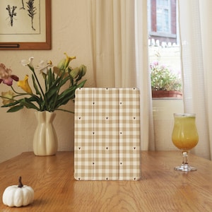 May include: A tan and white checkered tablet case with small black heart accents. The case is standing upright on a wooden table. A white pumpkin, a vase of flowers, and a glass of juice are also on the table.