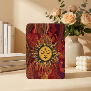 May include: A tablet case with a sun design, featuring a smiling face and yellow and orange rays. The background has red and brown patterns. The case is on a beige surface, with books and flowers in the background.
