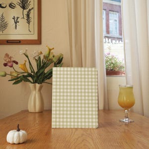Può includere: Un quaderno a scacchi verde chiaro e bianco è appoggiato su un tavolo di legno. Una zucca bianca, un vaso di fiori e un bicchiere di succo d'arancia sono anche sul tavolo. Una stampa botanica incorniciata è appesa al muro.
