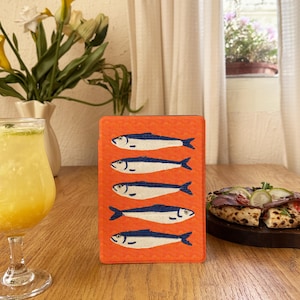 Puede incluir: Una funda naranja con un patrón repetido de cinco sardinas estilizadas. Cada sardina está representada en blanco con detalles azules. La funda está sobre una mesa de madera. El diseño es sencillo y llamativo.
