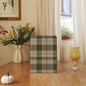 May include: A rectangular tablet case with a green and beige plaid pattern. The case is standing upright on a wooden table. A white pumpkin, a vase of flowers, and a glass of orange liquid are also on the table.