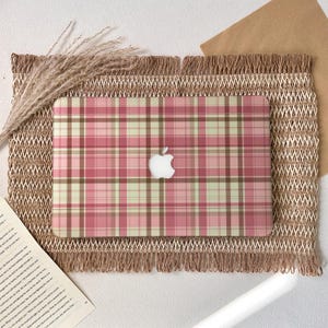 Könnte beinhalten: Eine Laptop-Hülle mit einem rosa, beigen und braunen Karomuster. Die Hülle hat das Apple-Logo in Weiß. Sie liegt auf einer gewebten, gefransten Tischdecke mit getrockneten Weizenähren.