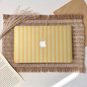 Può includere: Una custodia per laptop a righe gialle e crema con il logo Apple. La custodia è su un sottobicchiere intrecciato con un bordo a frange. Steli di grano essiccati sono a sinistra del laptop.