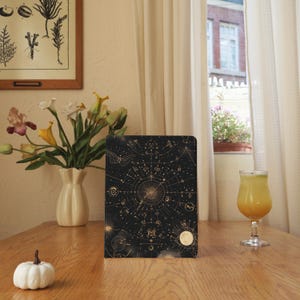 May include: A black notebook with a gold celestial design featuring constellations and astrological symbols. The notebook is on a wooden table, next to a vase of flowers and a small white pumpkin. A glass of orange juice sits nearby.