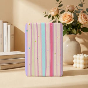 May include: A rectangular e-reader case featuring a vertical striped pattern in shades of light pink, cream, and turquoise, accented with small scattered multicolored dots. The case has a solid light purple spine and a textured surface finish.