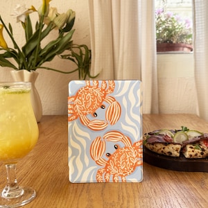 Puede incluir: Una funda rectangular con dos ilustraciones de cangrejos naranjas sobre un fondo azul claro con patrones de olas blancas. Los cangrejos tienen diseños detallados. La funda está sobre una mesa de madera.