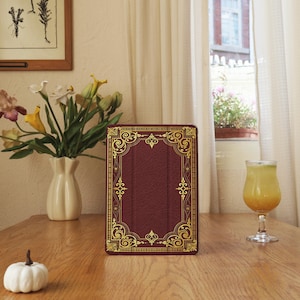 Pode incluir: Uma capa de tablet bordô com detalhes em filigrana dourada, assemelhando-se a uma capa de livro antigo. A capa está sobre uma mesa de madeira, ao lado de um vaso de flores e um copo de suco de laranja. O design é ornamentado e elegante.