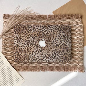 May include: A laptop with a leopard print cover featuring the Apple logo, resting on a woven mat with fringe. The mat is a light brown color, and the background is white. A sprig of dried grass is on the left.