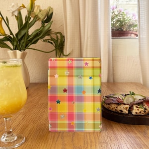 Pode incluir: Uma capa retangular para tablet com um padrão xadrez multicolorido vibrante em tons de amarelo, rosa, verde e azul, decorada com inúmeros pequenos ícones em forma de estrela espalhados em várias cores por toda a superfície.