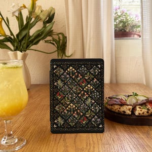 May include: A black e-reader case featuring a repeating diamond pattern filled with colourful botanical illustrations of flowers, leaves, and small birds, surrounded by a delicate vine border on a dark background.