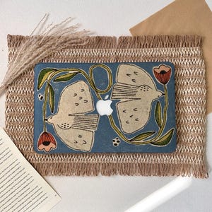 May include: A blue laptop skin featuring a printed illustration of two beige birds facing each other, surrounded by green vines and red flowers. The design includes a central cutout for the Apple logo, showcasing a textured, artistic aesthetic.