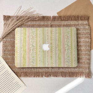 Può includere: Un computer portatile con una cover decorativa a righe pastello verdi, rosa e beige. Il logo Apple è visibile. Il portatile è posizionato su un tappetino strutturato con frange.