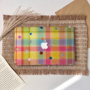 Può includere: Una custodia per laptop con un motivo a quadri colorato nei toni del giallo, rosa, verde e blu, adornata con decorazioni a forma di stella. Il logo Apple è visibile. La custodia è posizionata su una superficie tessuta e strutturata.