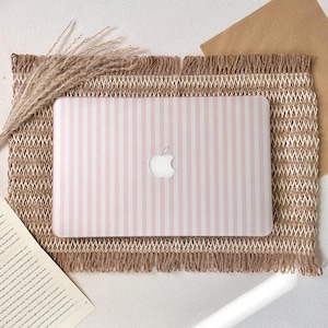 Puede incluir: Una funda para portátil a rayas rosas y blancas con el logotipo de Apple está centrada en una estera marrón tejida y texturizada con un borde con flecos. Espigas de trigo secas y un trozo de papel en el fondo.