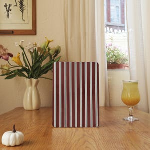May include: A notebook with burgundy and white vertical stripes stands on a wooden table. A white pumpkin and a vase of flowers are also on the table. A glass of orange juice is to the right of the notebook.