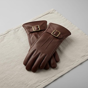 May include: A pair of brown leather gloves with a textured finish. Each glove features a strap with a gold-colored buckle. The gloves are resting on a light-colored, textured surface.
