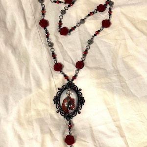 Op de afbeelding: Een lange ketting met rode rozenaccenten en zwarte kralen. De hanger is een ovaal frame met een portret. De ketting heeft een gotische of Victoriaanse uitstraling.