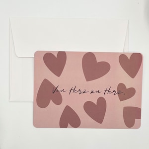 Könnte beinhalten: Eine Grußkarte mit rosa Hintergrund und mehreren weinroten Herzen. Die Karte trägt den deutschen Text "Von Herz zu Herz" in einer handschriftlichen Schriftart. Ein weißer Umschlag befindet sich hinter der Karte.