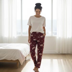 May include: Burgundy pajama pants with a white Dr. Pepper logo pattern are paired with a white t-shirt. The pants are loose-fitting and the t-shirt is short-sleeved. The person is standing in a well-lit room with a bed and window.