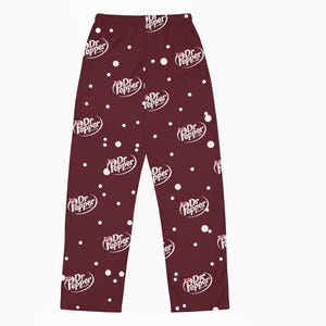 May include: Burgundy pajama pants with a repeating pattern of the Dr Pepper logo and white polka dots. The Dr Pepper logo is in white with a red bow. The pants are made of a soft material.