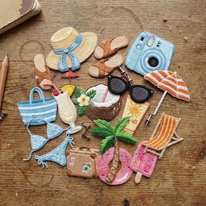 May include: Embroidered beach-themed items including a straw hat with a blue ribbon, sunglasses, a camera, a beach bag, a bikini, a cocktail, a coconut, a tube of sunscreen, a palm tree, a suitcase, a donut float, and a popsicle.