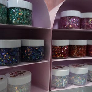 May include: Multiple small clear plastic jars filled with various colors of chunky glitter, including green, blue, red, and iridescent white. The jars feature white screw-top lids and are organized on shelves within two light pink arched displays.