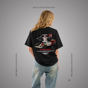 May include: Black t-shirt featuring a graphic of a McLaren race car with the text "AYRTON SENNA" and "McLaren MP4/4". The shirt also has the text "MASTERWORKSNY" and "MCLAREN 26 COLLECTION".