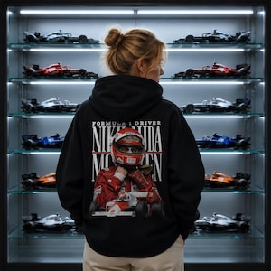 Può includere: Una felpa nera con cappuccio e stampa grafica sul retro. Il design mostra un pilota di Formula 1 con tuta e casco rossi sopra un'auto da corsa. Il testo recita FORMULA 1 DRIVER NIKI LAUDA MUNCHEN.