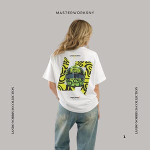 Könnte beinhalten: Weißes T-Shirt mit einer Grafik eines Rennhelms, dem Text "LANDO NORRIS" und der Nummer "26". Das Design beinhaltet ein gelb-schwarzes Muster. Das Shirt zeigt auch "MASTERWORKSNY" und "LANDO NORRIS 26 COLLECTION".