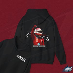 Może przedstawiać: Czarna bluza z kapturem z grafiką Charlesa Leclerca w kombinezonie wyścigowym, z obrysem toru Grand Prix Monako. Bluza ma również napis "CHARLES LECLERC" i wzór w szachownicę.
