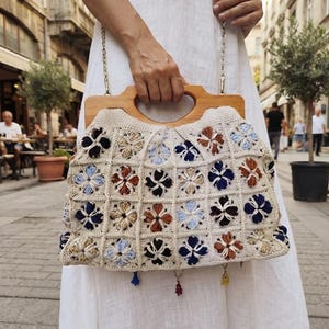 May include: A crochet handbag featuring a grid of square floral motifs in shades of blue, brown, and cream. The bag has a light-toned wooden handle with a cutout grip and a thin metal chain strap. Small colorful charms hang from the bottom edge.