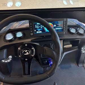 May include: Black golf cart interior with a perforated leather steering wheel. The dashboard features a digital display, gauges, and a speaker. The steering wheel has a black center cap with a white logo.