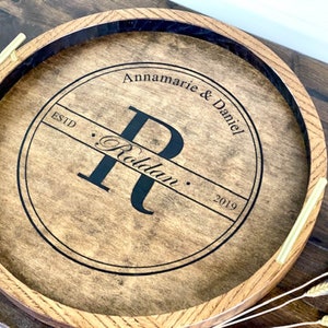 Puede incluir: Una bandeja redonda de madera con un c&iacute;rculo negro en el centro. El c&iacute;rculo tiene el texto "Annamarie & Daniel" y "Estd. Roldaun. 2019" y una letra "R" grande en el centro. La bandeja tiene dos asas doradas.