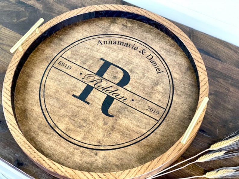 Puede incluir: Una bandeja redonda de madera con un c&iacute;rculo negro en el centro. El c&iacute;rculo tiene el texto "Annamarie & Daniel" y "Estd. Roldaun. 2019" y una letra "R" grande en el centro. La bandeja tiene dos asas doradas.