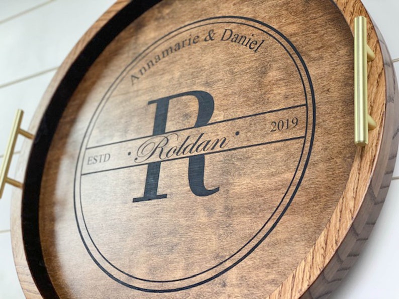 Puede incluir: Una bandeja redonda de madera con asas de metal dorado. La bandeja tiene un c&iacute;rculo negro en el centro con el texto "Annamarie & Daniel" y "ESTD. Roldan. 2019" en una fuente cursiva.