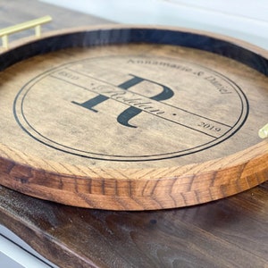 Puede incluir: Una bandeja redonda de servir de madera con asas de metal dorado. La bandeja tiene un c&iacute;rculo negro en el centro con el texto "Annamarie & Daniel" y la letra "R" en una fuente de escritura. El texto "Est. 2019" tambi&eacute;n est&aacute; incluido.