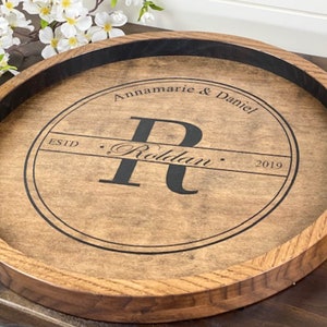 Puede incluir: Una bandeja redonda de madera para servir con asas de lat&oacute;n. La bandeja tiene un borde negro y un dise&ntilde;o circular en el centro que dice "Annamarie & Daniel Estd. - Rolldan - 2019" con una gran "R" en el centro.