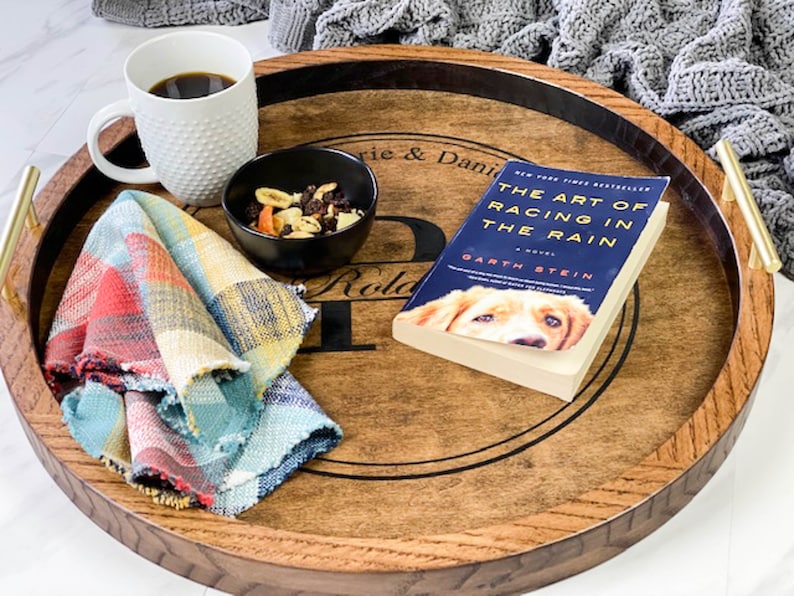 Puede incluir: Una bandeja de madera con un borde negro y asas doradas. La bandeja tiene una inscripci&oacute;n personalizada que dice "Laurie & Daniel" y "Roland" con una gran "P" en el medio. La bandeja est&aacute; llena de un libro, una taza de caf&eacute;, un taz&oacute;n de mezcla de frutos secos y una servilleta de tela a cuadros.