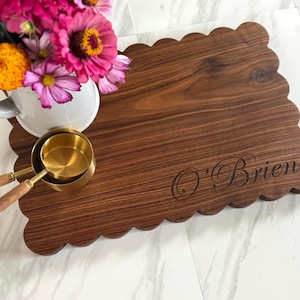 Könnte beinhalten: Ein Servierbrett aus Walnussholz mit gewelltem Rand, graviert mit dem Namen "O'Brien". Das Brett befindet sich neben einer weißen Tasse mit rosa und orangefarbenen Blumen und zwei Messbechern aus Messing.
