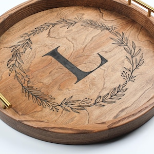 May include: Round wooden serving tray with a dark brown rim and gold handles. The tray features a light brown surface with a black floral wreath design and a large black letter "L" in the center.