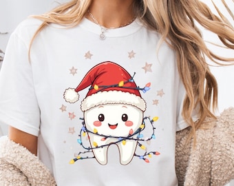 May include: White t-shirt featuring a cartoon tooth wearing a red Santa hat with a white trim. The tooth is smiling and wrapped in a string of colorful Christmas lights. Small star shapes are scattered around the design.