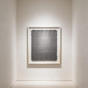 May include: A framed artwork with a gray, textured surface in a herringbone pattern. The artwork is displayed in a white-walled gallery space. The frame is a light wood color.