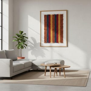 May include: A framed artwork featuring a textile with vertical stripes in red, yellow, and blue. The artwork is displayed on a white wall in a living room setting. The room includes a gray sofa, wooden coffee tables, and a potted plant.