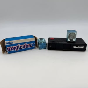 May include: A selection of vintage photographic equipment. The image shows a blue and white "magicube" box, two "magicubes", and a black "Halina" camera. The "magicube" box is open. The camera has a red button and the brand name "Halina".