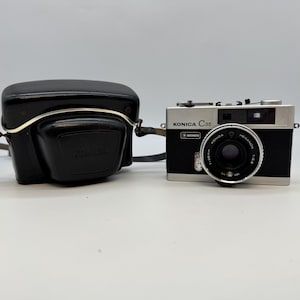 May include: A vintage Konica C35 camera with a black and silver body, and a black leather case. The camera has a Hexanon 1:2.8 lens and the word "Konica" is visible on the lens and body. The case is black with the Konica logo.